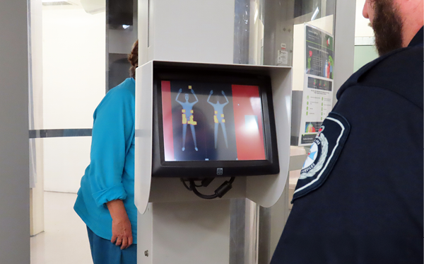 Women standing in body scanner while a correctional officer inspects a read-out screen.