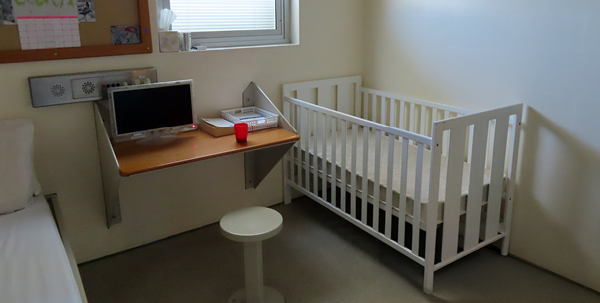 View of room with a single bed along one wall and a cot on the other wall, a small table and chair sits under a noticeboard and window.