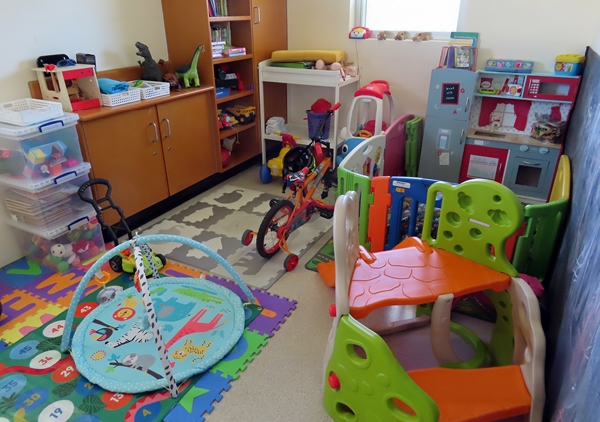 A small room stocked with many items for small children including a change table, baby play-mat, small slide, play kitchen, tricycle and a selection of toys.